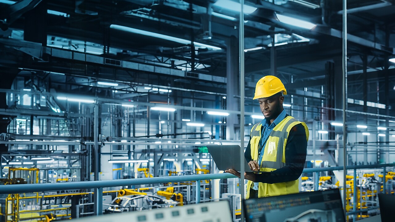 A male engineer wearing a yellow hard hat and high-visibility vest stands in a modern, high-tech automotive factory. 