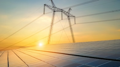 A field of solar panels with a high-voltage transmission tower and power lines in the background, set against a warm sunrise sky.
