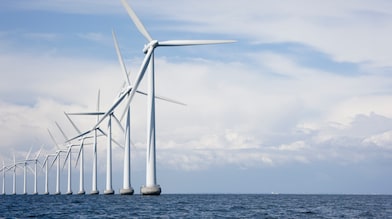A row of white offshore wind turbines stretching into the distance, installed in the ocean under a partly cloudy sky.