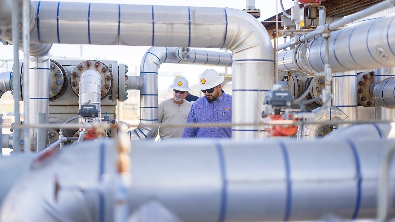 two people stand facing each other conversing. They are stood in an industrial plant with metal pipes