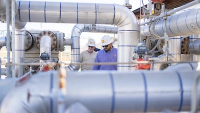 two people stand facing each other conversing. They are stood in an industrial plant with metal pipes