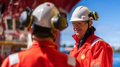 Two males in Shell branded protective workwear are stood opposite eachother talking. The male in the foreground has his back to the camera, the second male is facing the camera