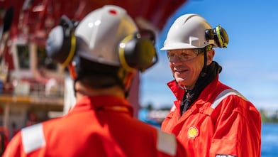 Two males in Shell branded protective workwear are stood opposite eachother talking. The male in the foreground has his back to the camera, the second male is facing the camera