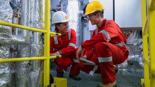Two colleagues in Shell branded protective work wear are crouched down on the floor of a process container as if monitoring and discussing the apparatus they are crouched down in front of