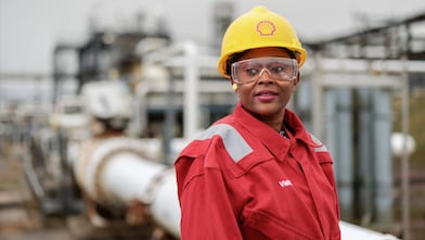 Image of a female wearing Shell branded protective workwear in the foreground standing in front of a blurred background of a processing plant.