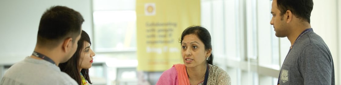 A mixed group of Indian employees are gathered around a table, listening to a senior female staff. 
