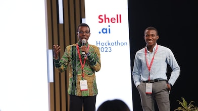 two men at a stage, speaking to a microphone, Shell.ai logo in the background