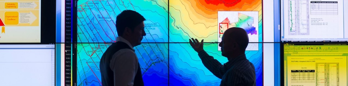 Two men are standing in front of a wall-sized screen and are discussing. The screen displays an unidentified topographic visualisation and other smaller data in the form of tables. On man is pointing at the screen as if sustaining his point with the data displayed.