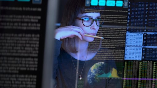 Person wearing glasses analysing data on a computer screen, with digital charts and data visuals overlaid in the background.