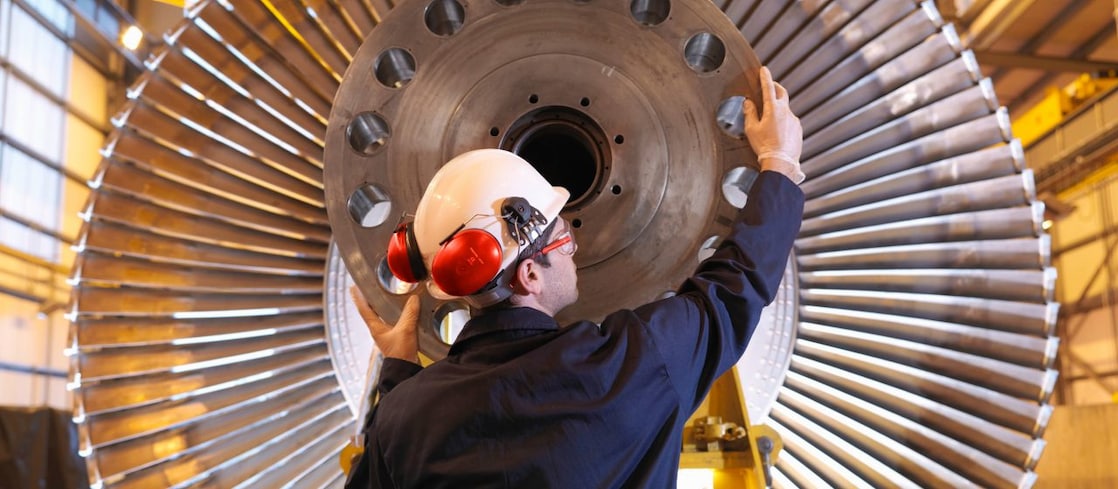 Person wearing a hard hat and ear protection inspecting a large industrial turbine component inside a workshop.
