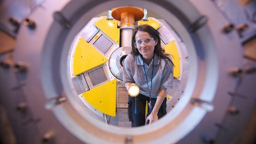 Person wearing protective eyewear examining industrial equipment through a circular aperture, using a handheld torch to inspect components.