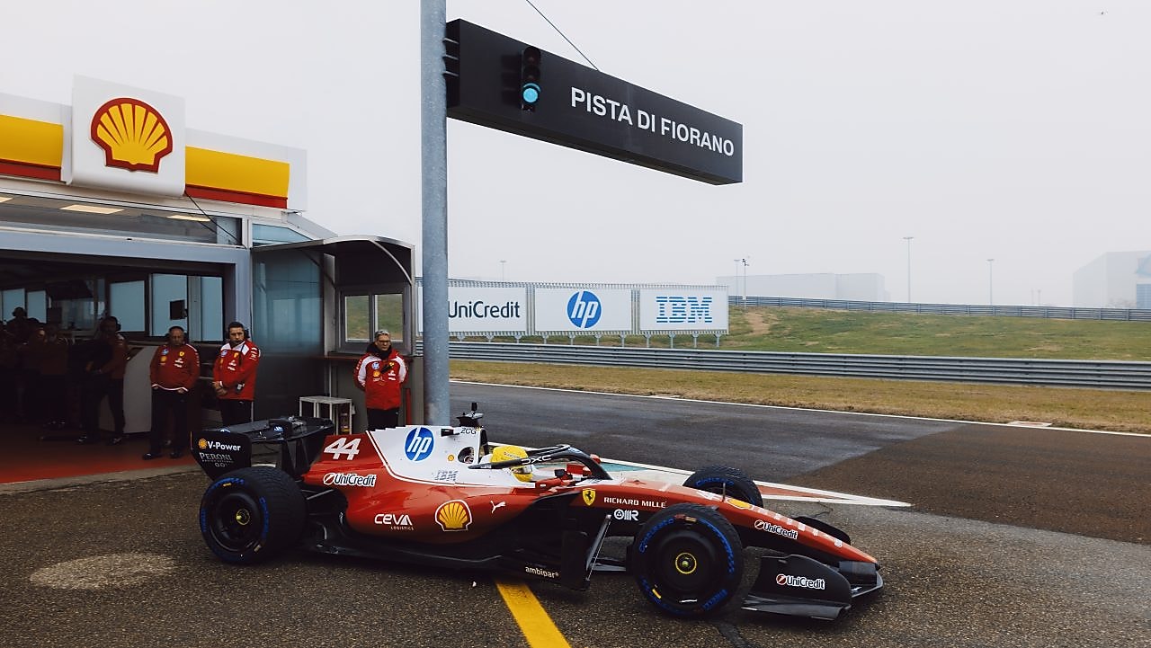 A Scuderia Ferrari HP Formula 1 car drives out of the garage at the Fiorano test track, with team members standing nearby.