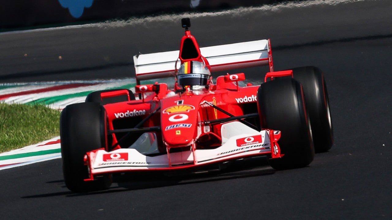 Ferrari F2002 car driving towards the camera on the Monza circuit, with the driver visible in the cockpit.