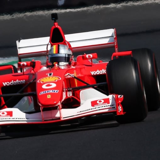 Ferrari F2002 car driving towards the camera on the Monza circuit, with the driver visible in the cockpit.