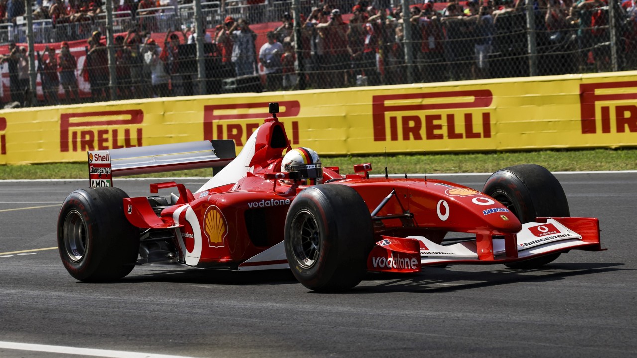 Ferrari F2002 car driving on the Monza circuit in front of trackside barriers and spectators.