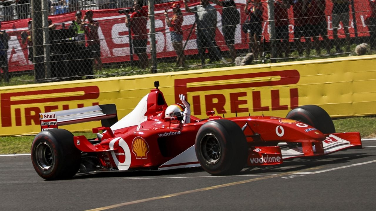 Side view of the Ferrari F2002 car on track at Monza, with the driver raising a hand towards spectators.