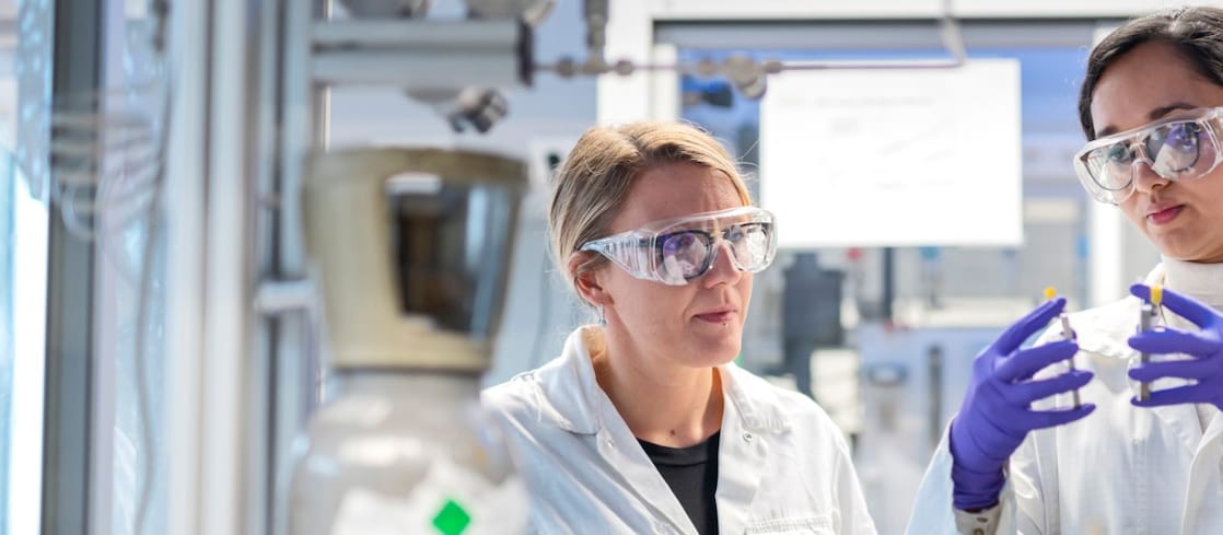 Two women look carefully to laboratory apparatus. They both wear protective glasses and gloves.