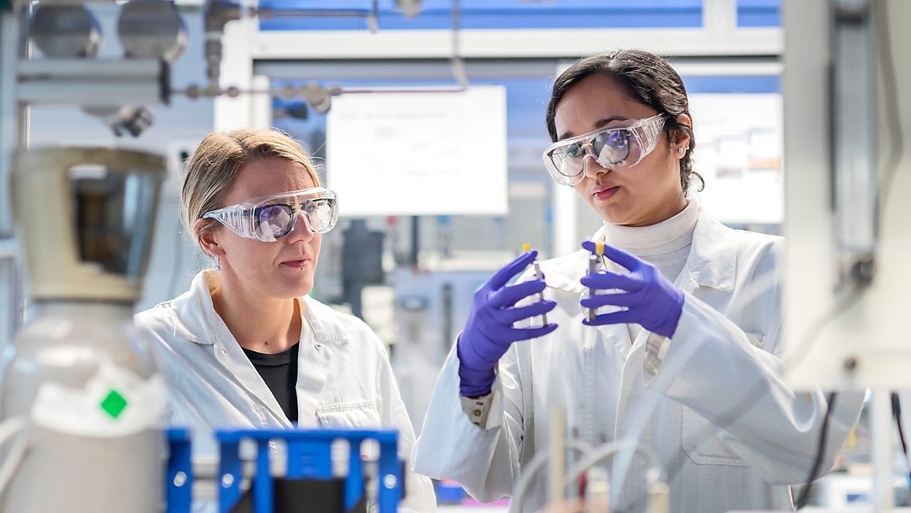 Two women look carefully to laboratory apparatus. They both wear protective glasses and gloves.