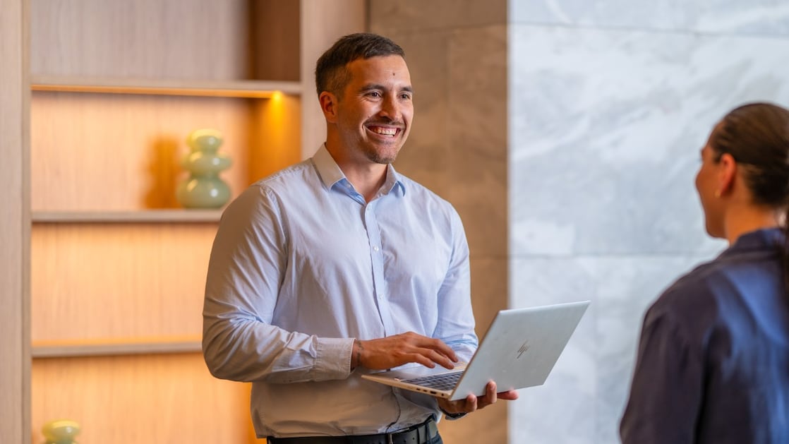 A person standing indoors, holding an open laptop and speaking with another individual. They are in a modern office space with wooden shelving and soft lighting in the background.
