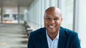 A person wearing a blue blazer and light-colored shirt is seated in a bright indoor corridor with large windows and a row of benches leading into the distance.