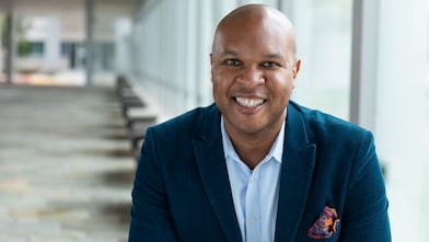 A person wearing a blue blazer and light-colored shirt is seated in a bright indoor corridor with large windows and a row of benches leading into the distance.
