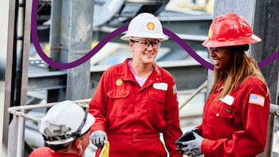 A worker wearing a white hard hat with a Shell logo and a red protective uniform stands in an industrial setting with metal structures in the background.