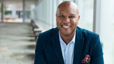 A person wearing a blue blazer and light-colored shirt is seated in a bright indoor corridor with large windows and a row of benches leading into the distance.