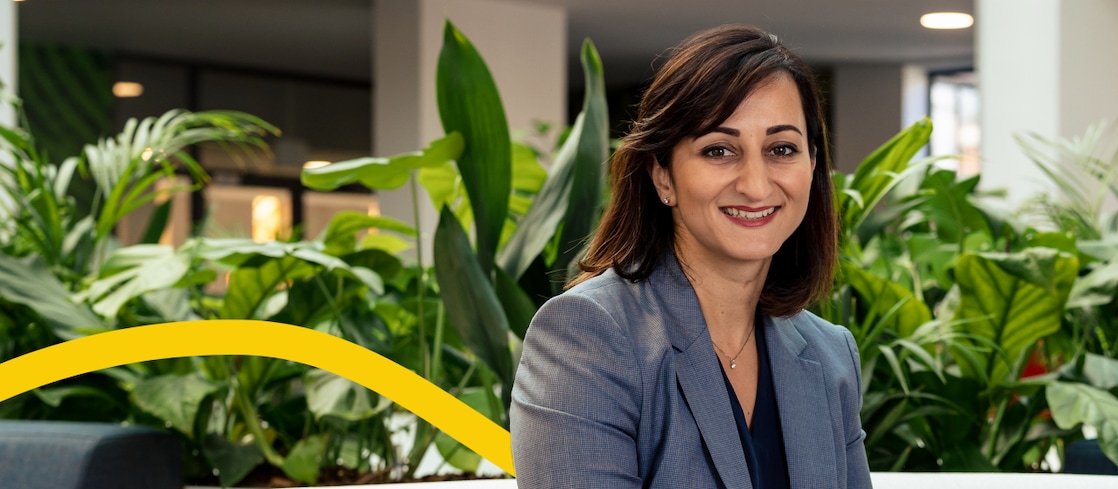 A lady in a grey blazer sits in an office environment among tall green plants, with a modern building visible in the background. 