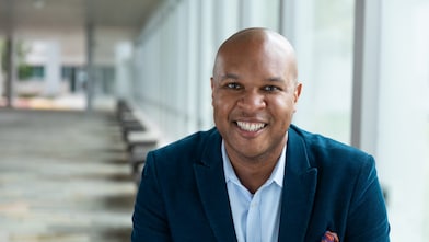 A person wearing a blue blazer and light-colored shirt is seated in a bright indoor corridor with large windows and a row of benches leading into the distance.
