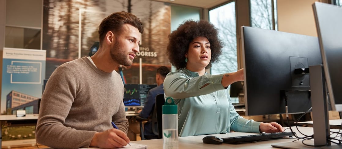 Two colleagues sit at a desk in a modern office. One takes notes while the other points at a large computer monitor, appearing to explain or demonstrate something. Office posters, windows, and other workers are visible in the background